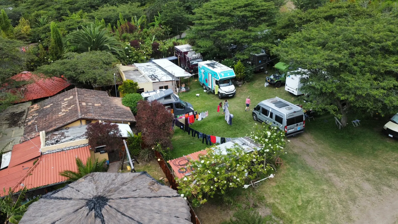 Vista aérea del camping: campers de overlanders estacionados entre los árboles, ropa tendida y los techos de teja de las cabañas